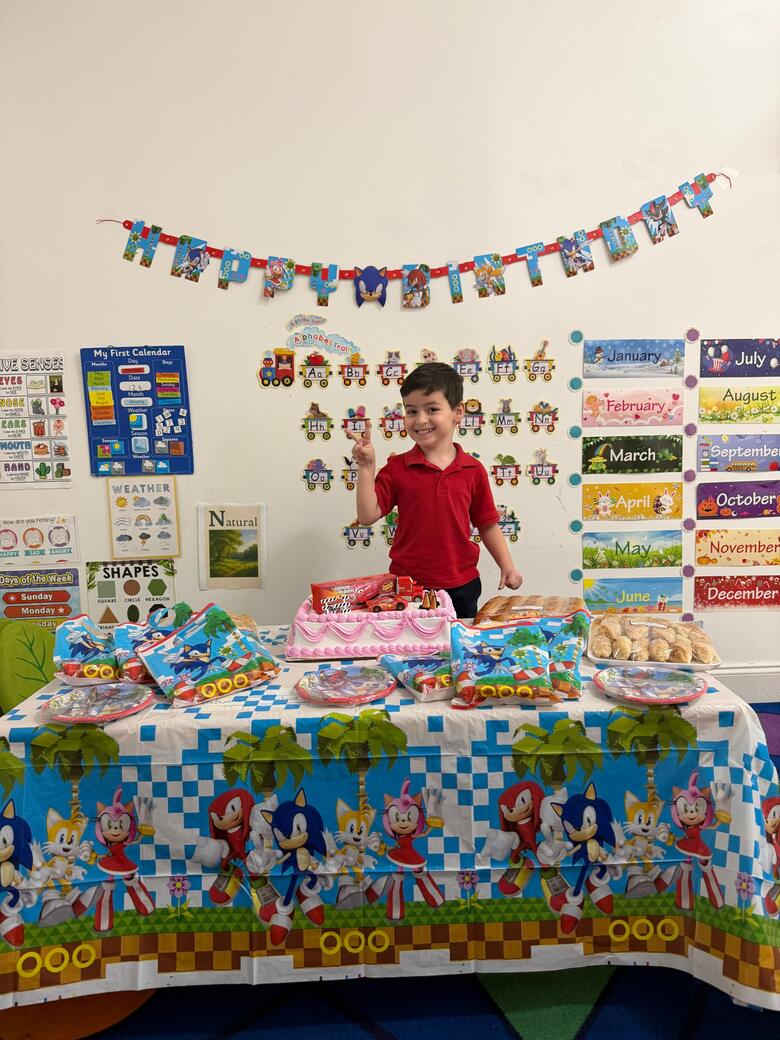 A boy standing behind a decorated table of cupcakes at a classroom birthday celebration.