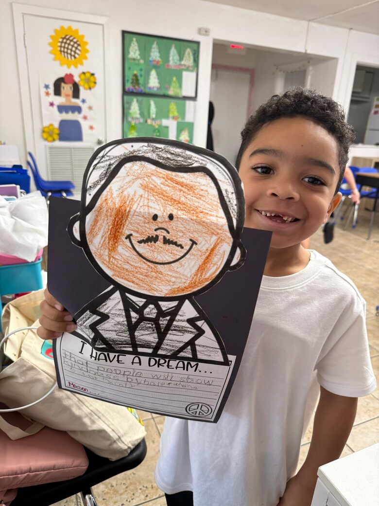 A boy grinning as he holds up his crayon drawing of Dr Martin Luther King Jr.