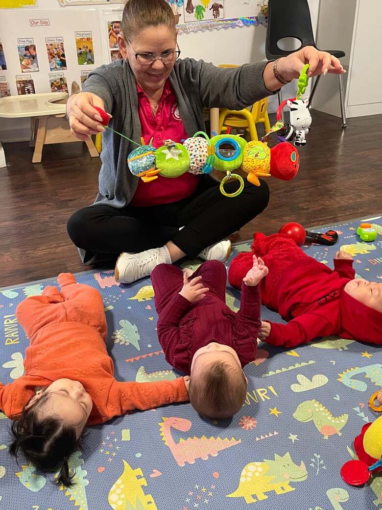 A teacher sitting cross-legged on a play mat, holding up a string of colourful rattles for three infants lying in front of her.