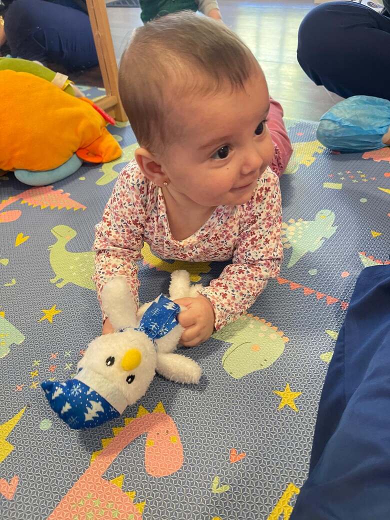 A baby propped up on her elbows on a patterned foam mat, holding a soft blue and white toy.