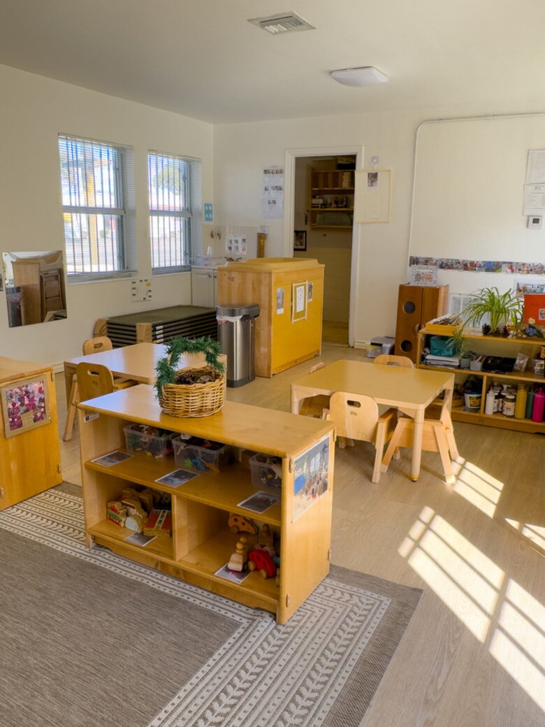 An infant room with pale wood floors, low wooden shelving and daylight coming through two windows.
