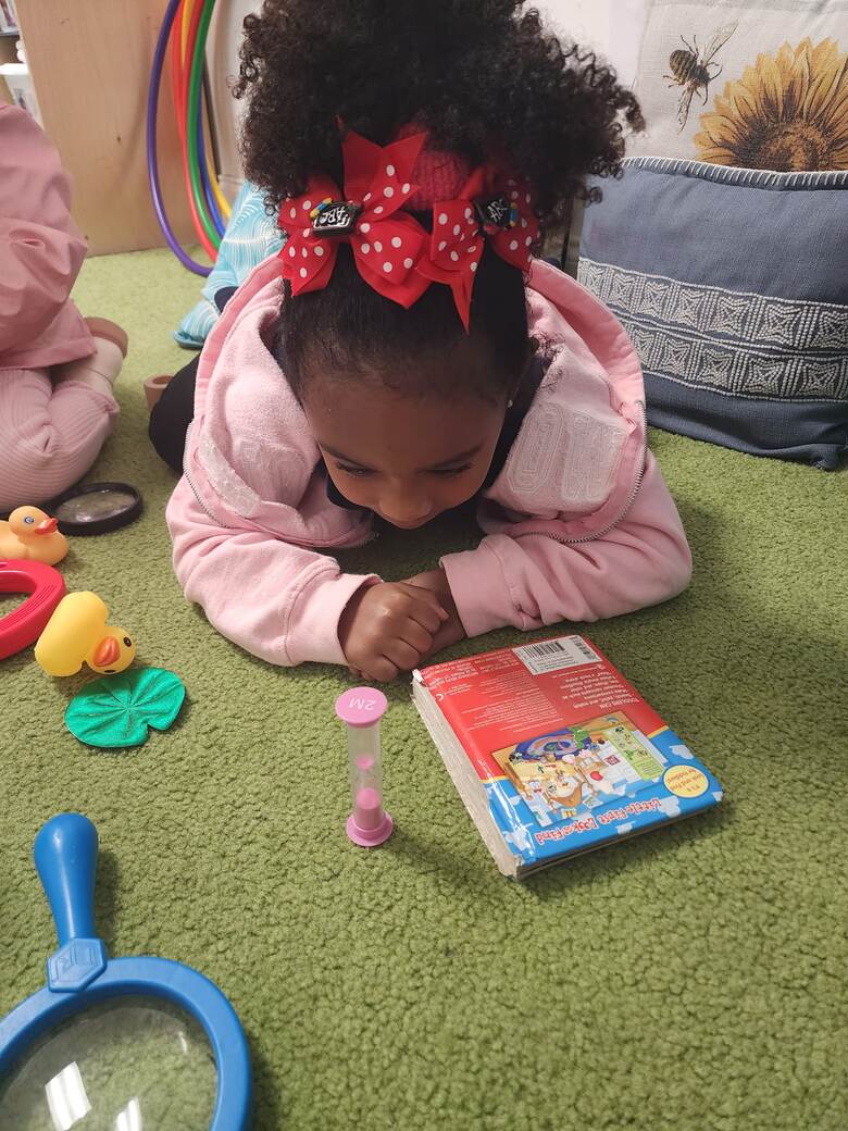 A girl lying on a green carpet with her chin on her hands, absorbed in a picture book.