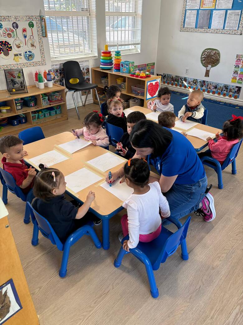 Children seated around a table on blue chairs, working on sheets of paper while a teacher helps.