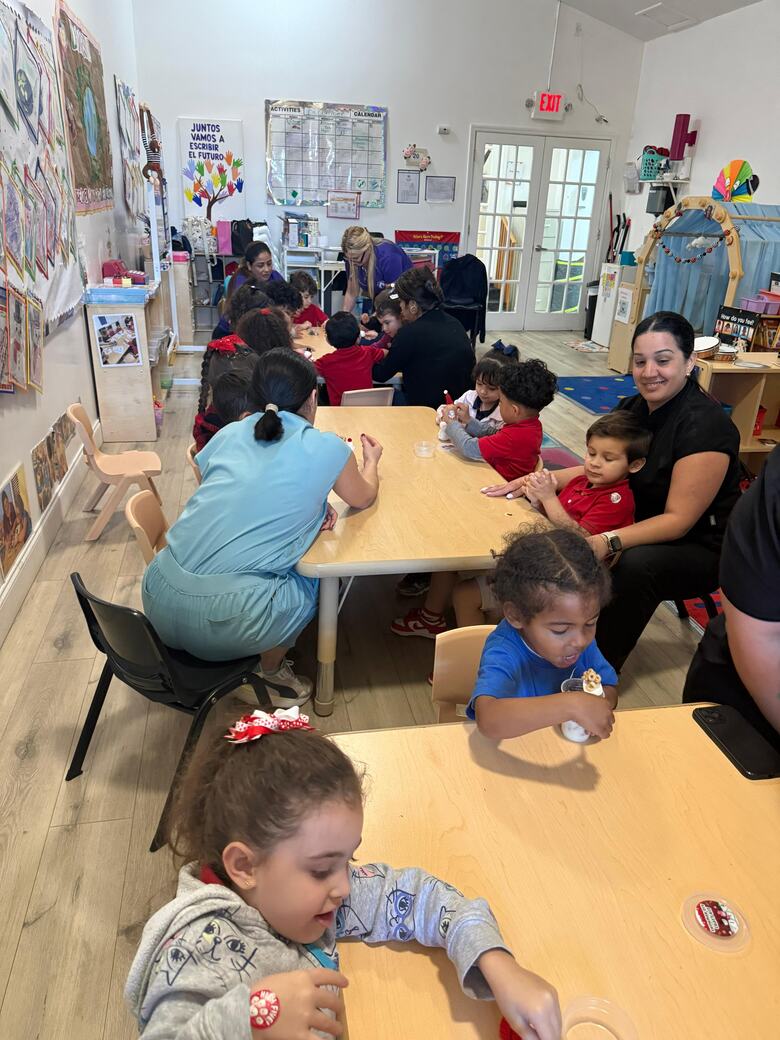 Toddlers seated at low tables in a bright classroom, a teacher leaning in to help.