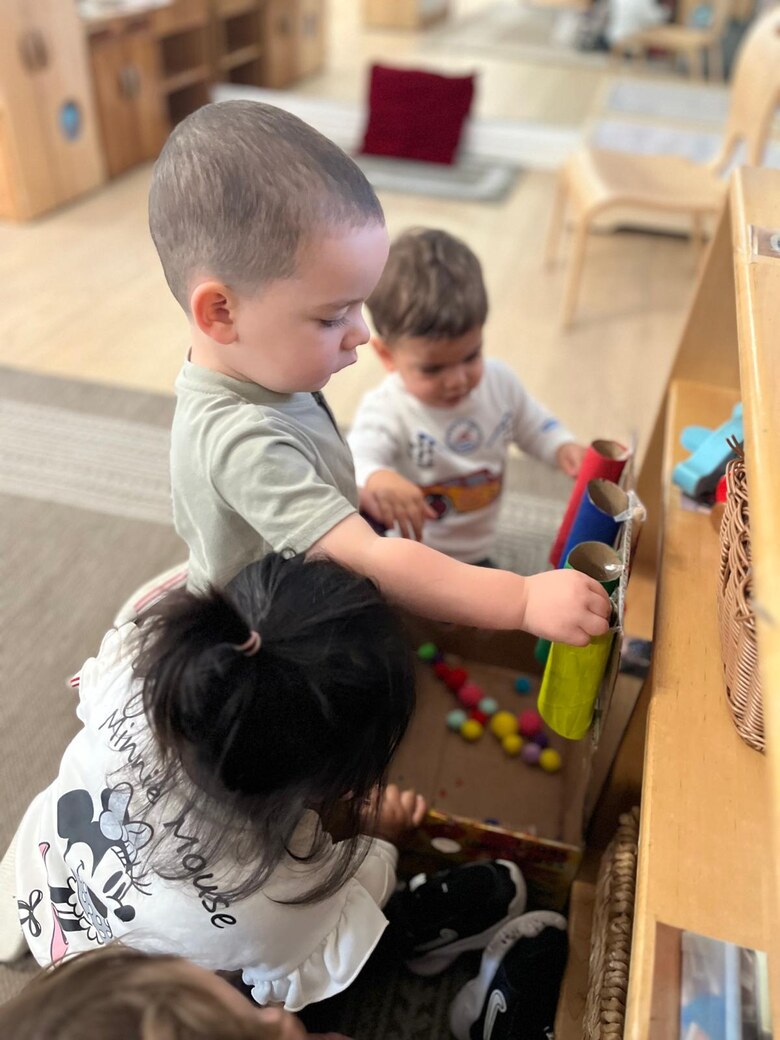 Two toddlers at a wooden shelf, posting coloured pompoms into cardboard tubes.