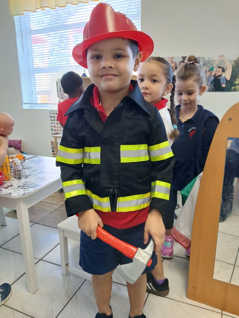 A boy in a firefighter's coat and red helmet holding a toy axe, classmates waiting behind him.