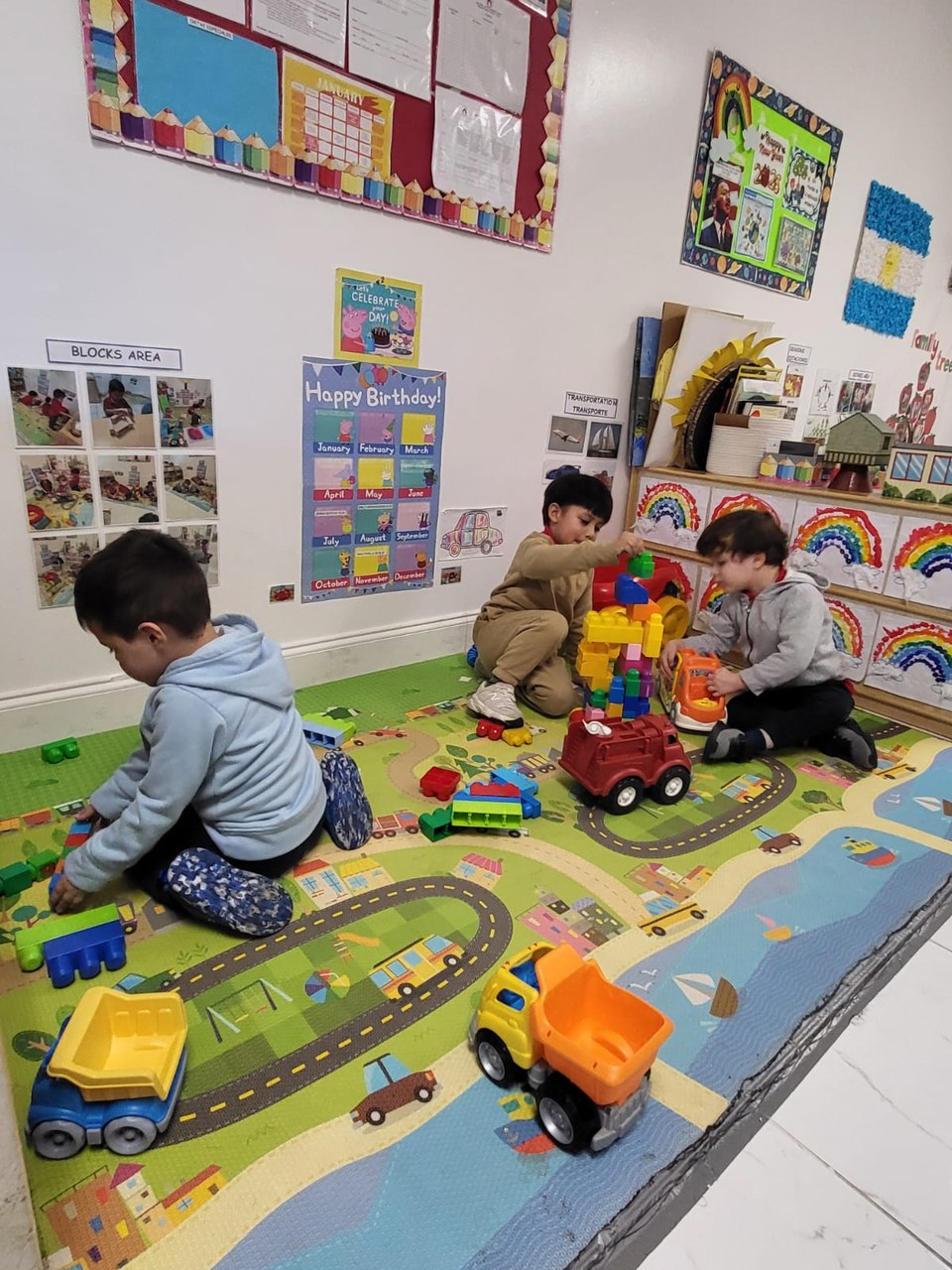 Three young children are engaged in play on a colorful mat featuring a road and buildings. They are building with blocks and interacting with toy vehicles. The classroom is decorated with educational posters and a calendar, suggesting a learning environment. The children appear focused and happy.