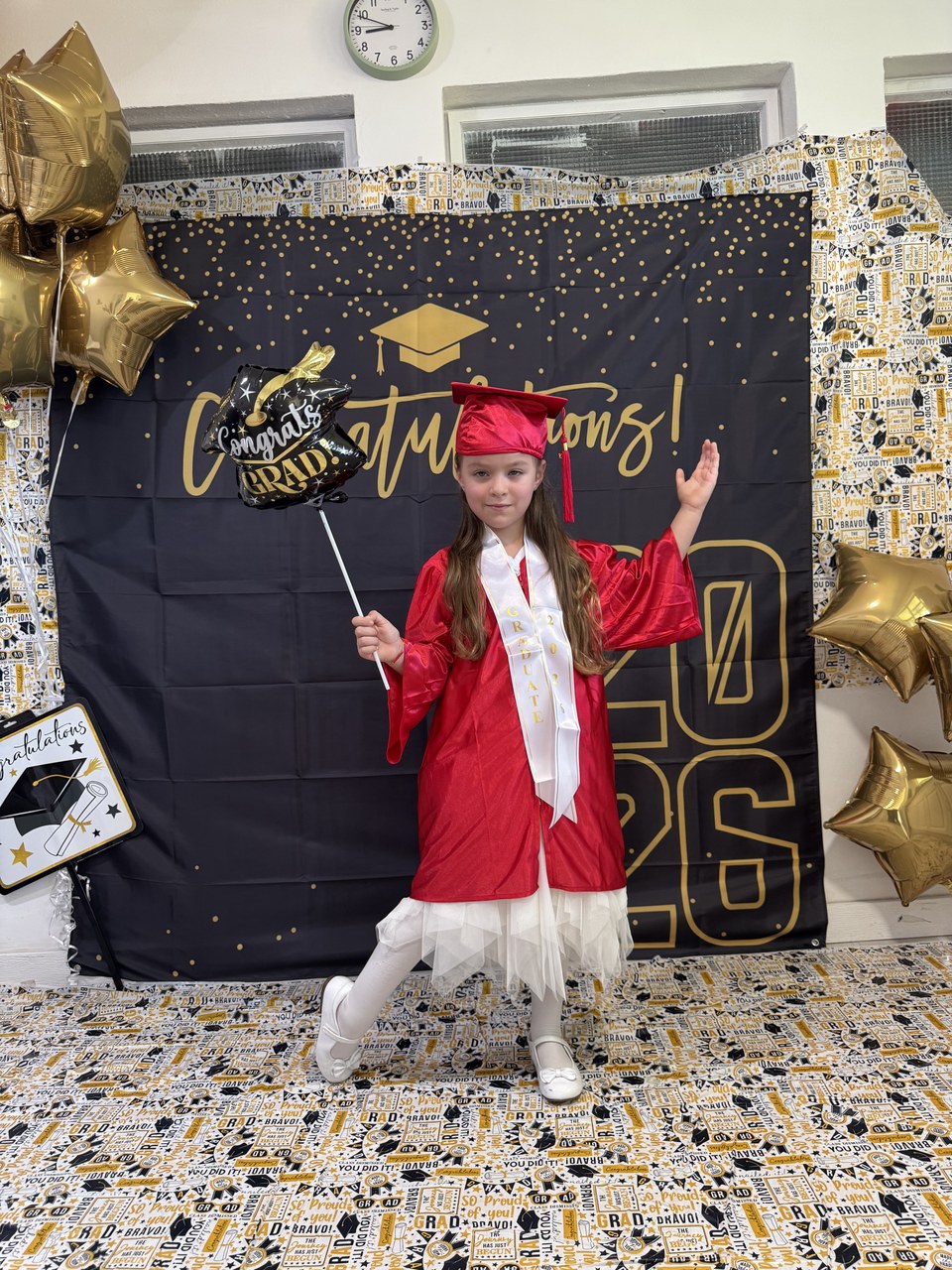 A young child in a red graduation gown and cap stands in front of a backdrop that reads "Congratulations!" with gold balloons and decorations.