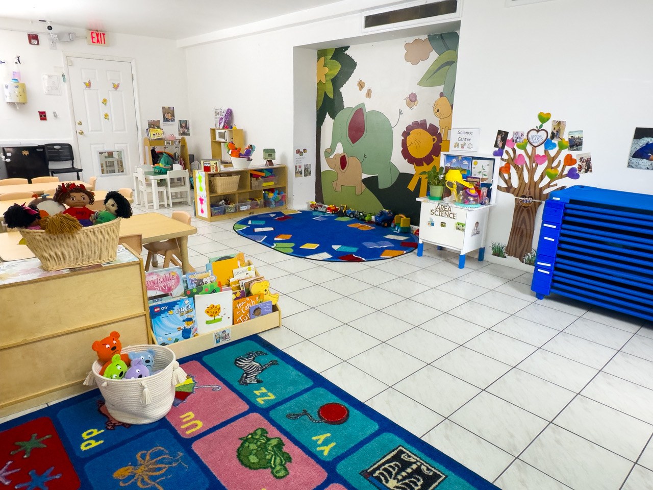 A classroom with a painted tree mural, an alphabet rug, and open toy shelves.