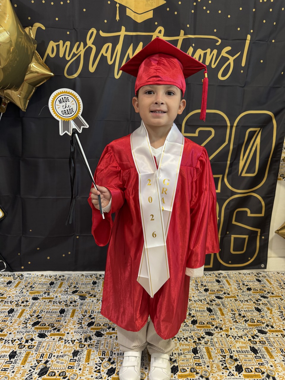 A young child stands proudly in a red graduation gown and cap, holding a diploma and a tassel, against a backdrop celebrating "Congratulations!".