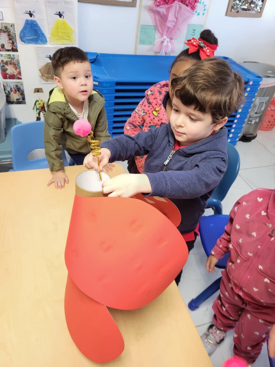 In a brightly lit classroom, three young children are engaged in a craft activity. One child, focused on a red paper sculpture, uses a stick to apply something inside it. Another child watches attentively, while a third child, partially obscured, appears to be assisting. The setting includes blue chairs, a wooden table, and colorful decorations on the walls, suggesting a creative and educational environment.