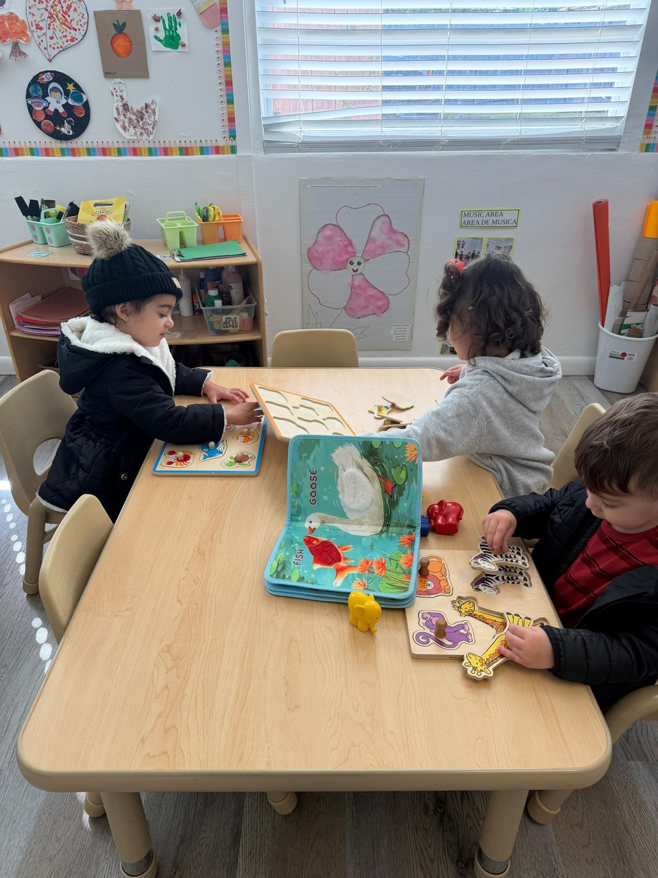 Three children are engaged in a quiet activity at a wooden table, surrounded by colorful educational materials including puzzles and books. The setting appears to be a classroom with shelves holding various supplies and decorations on the walls. The children seem focused and calm.