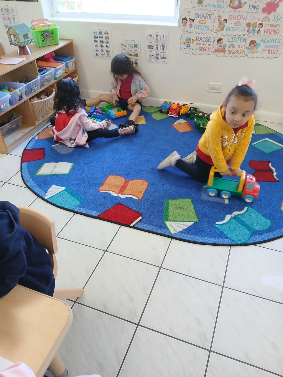 Three children are engaged in play on a colorful rug in a classroom setting. One child is building with blocks, another is interacting with toys, while the third is pushing a toy truck. The room features shelves with organized toys and books, and motivational posters adorn the walls. The setting appears to be during the daytime.